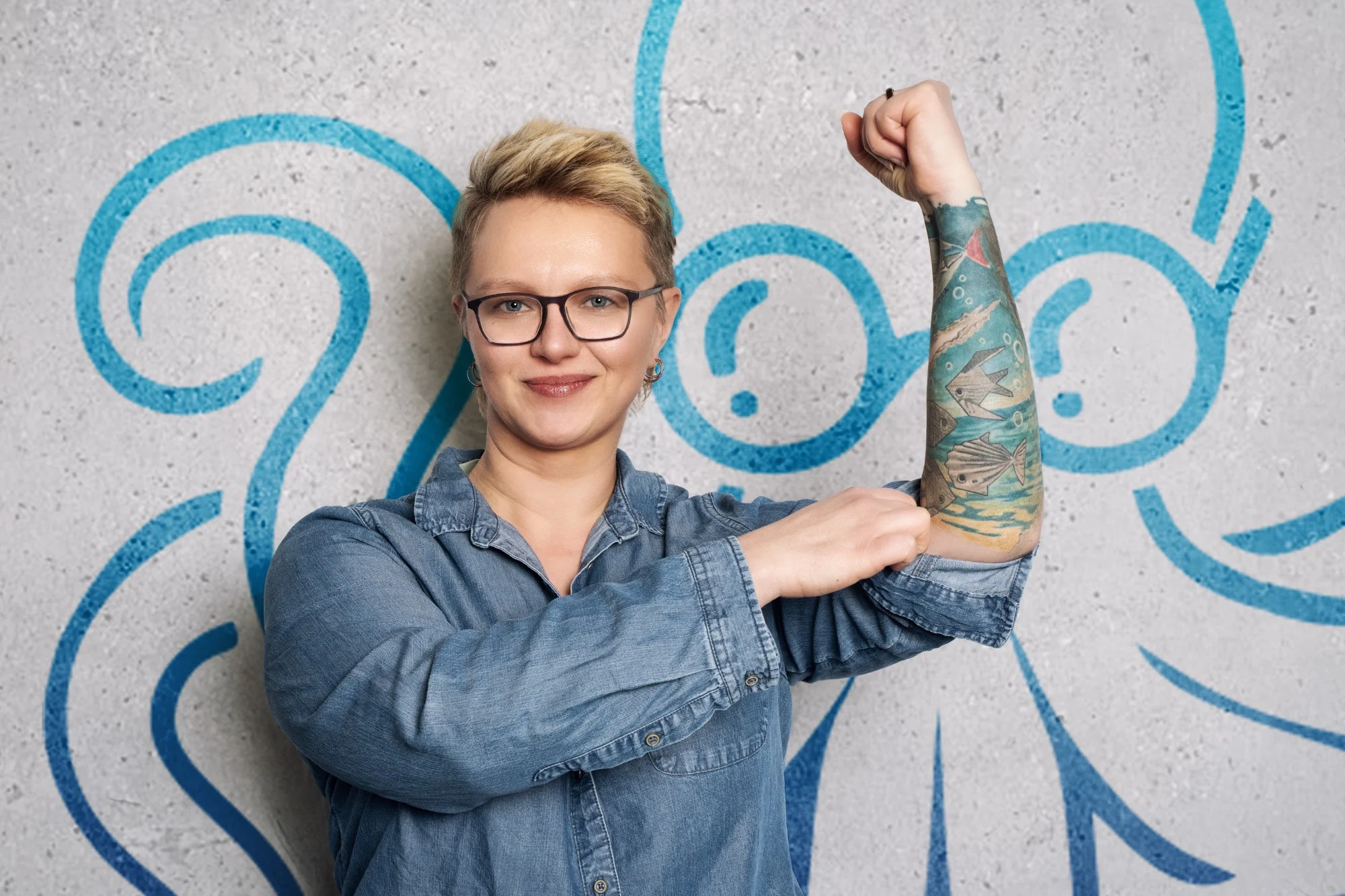 Anna Rachow steht vor einer Wand mit blauem Oktopus-Wandbild und zeigt lächelnd ihren angewinkelten Arm